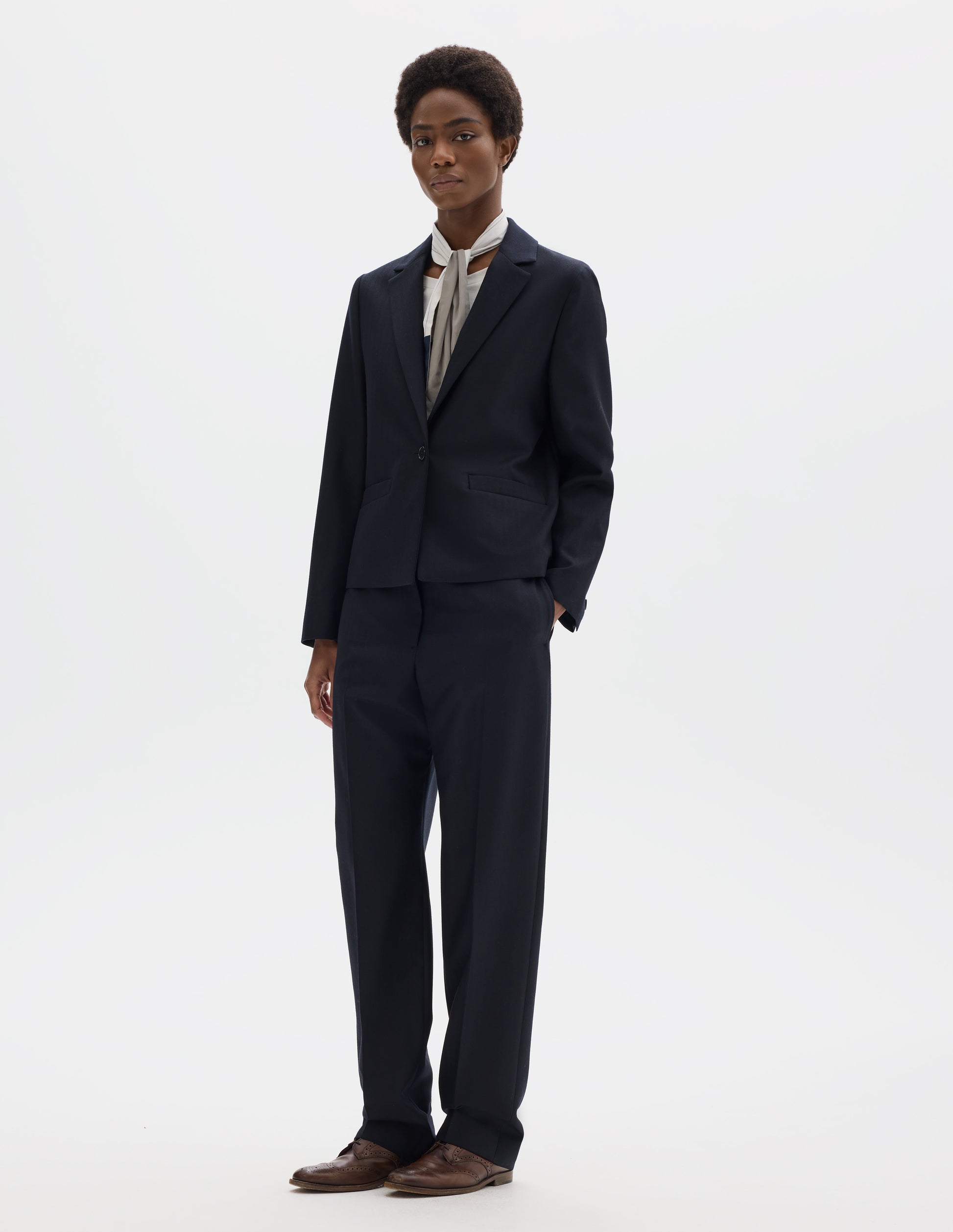 Full body image, front view of model wearing dark blue wool blazer with single horn button fastening, welt pockets and horn buttons on cuffs. Styled over white top, and with white and blue shirting scarf, dark blue matching trousers and brown leather brogues.