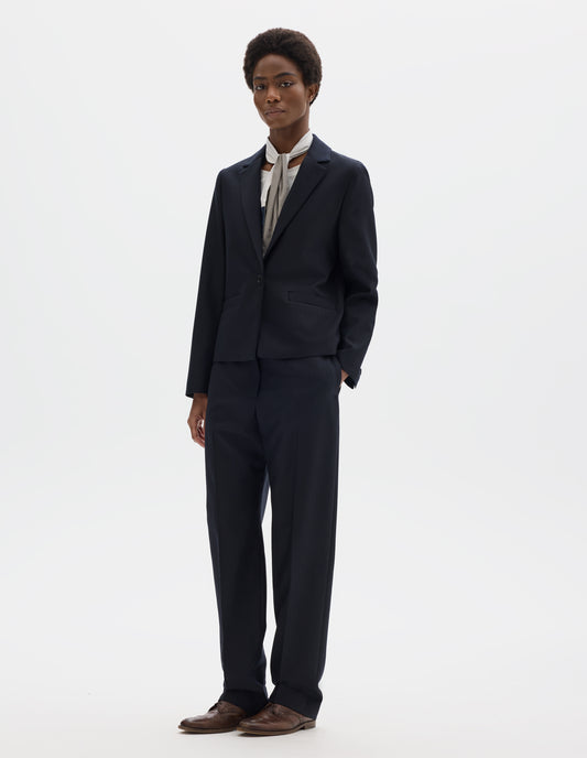 Full body image, front view of model wearing dark blue wool blazer with single horn button fastening, welt pockets and horn buttons on cuffs. Styled over white top, and with white and blue shirting scarf, dark blue matching trousers and brown leather brogues.
