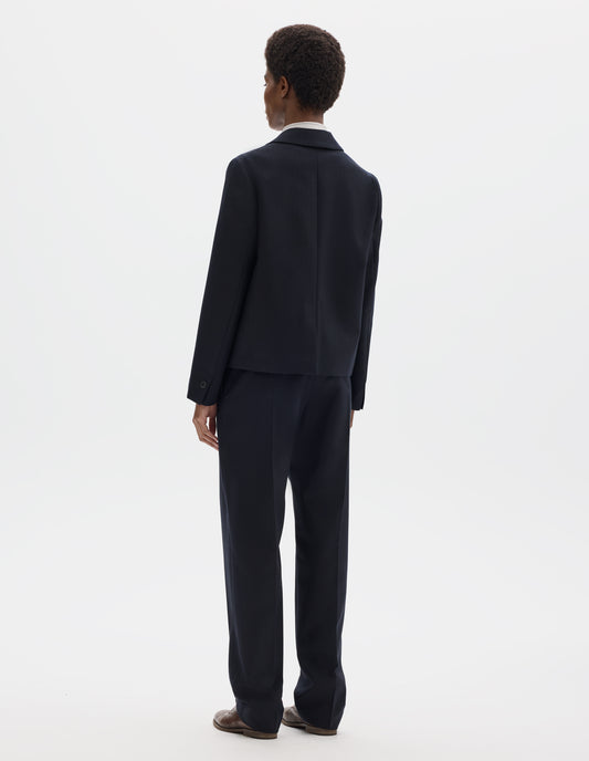 Full body image, back view of model wearing dark blue wool blazer with centre back vent and horn buttons on cuffs. Styled over white top, and with white and blue shirting scarf, dark blue matching trousers and brown leather brogues.