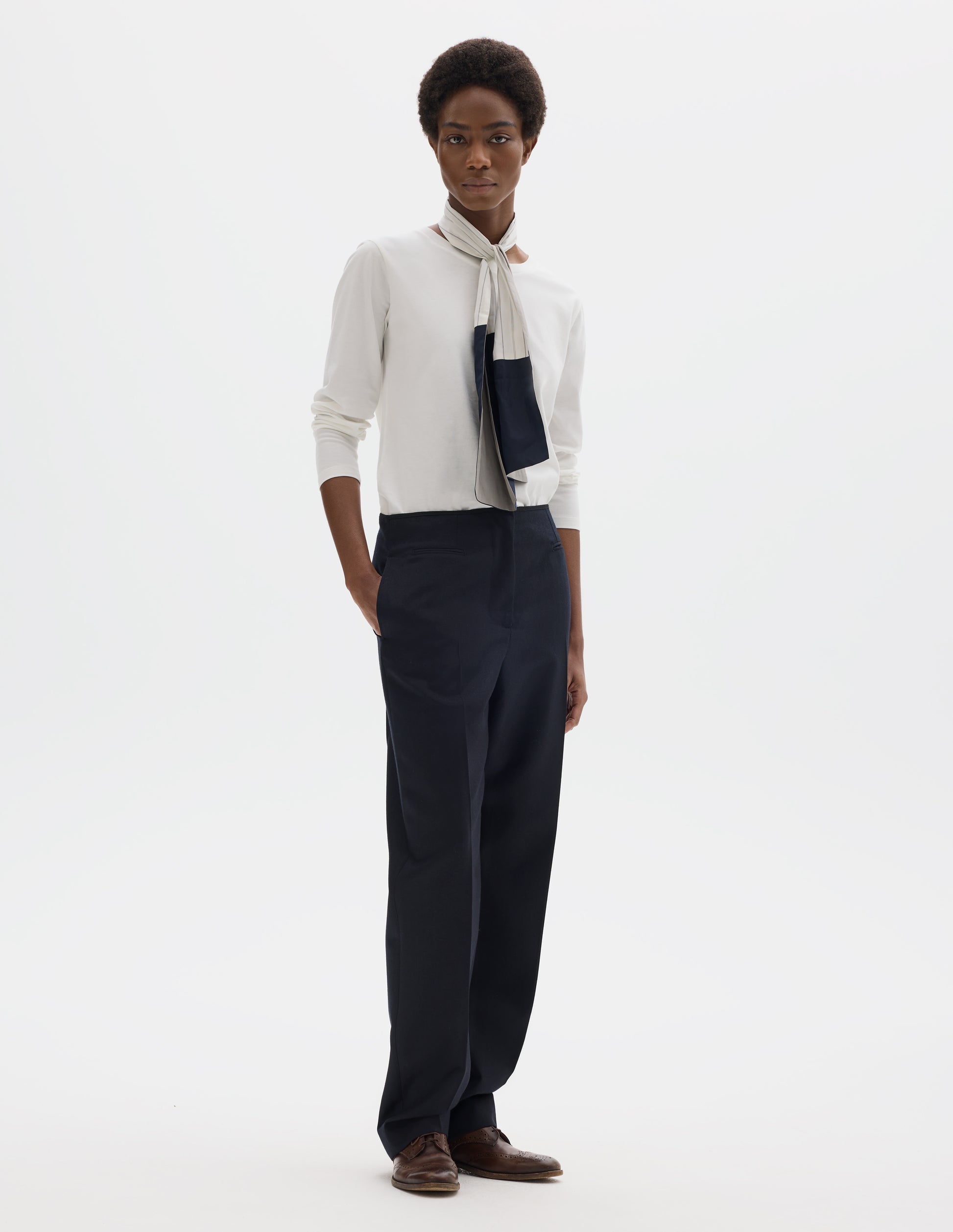 Full body image, front view of model wearing white relaxed cotton t-shirt with wide round neck, long sleeves and boxy cut. Styled with white and blue shirting scarf, dark blue trousers and brown leather brogues.