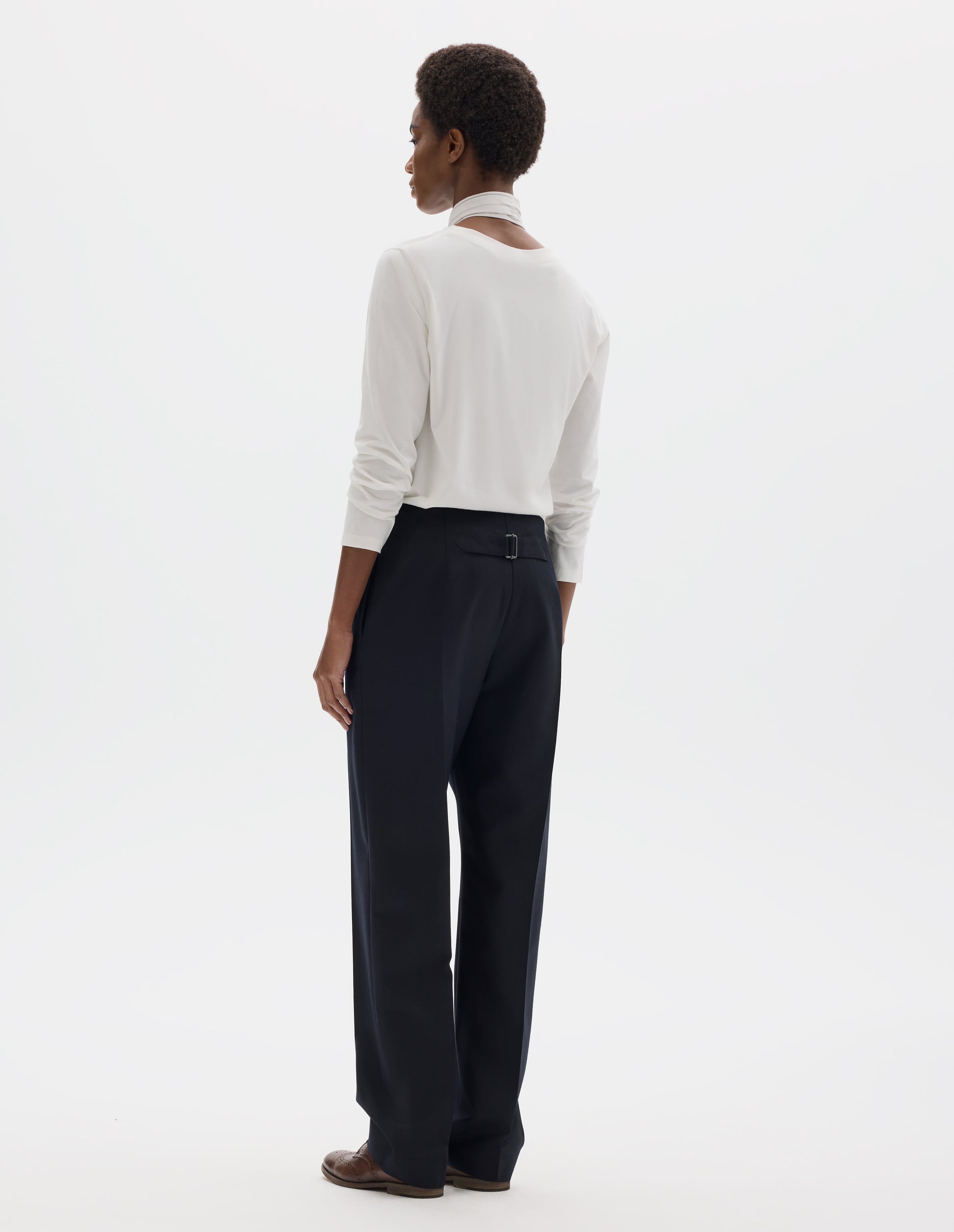 Full body image, back view of model wearing white relaxed cotton t-shirt with wide round neck, long sleeves and boxy cut. Styled with white and blue shirting scarf, dark blue trousers and brown leather brogues.