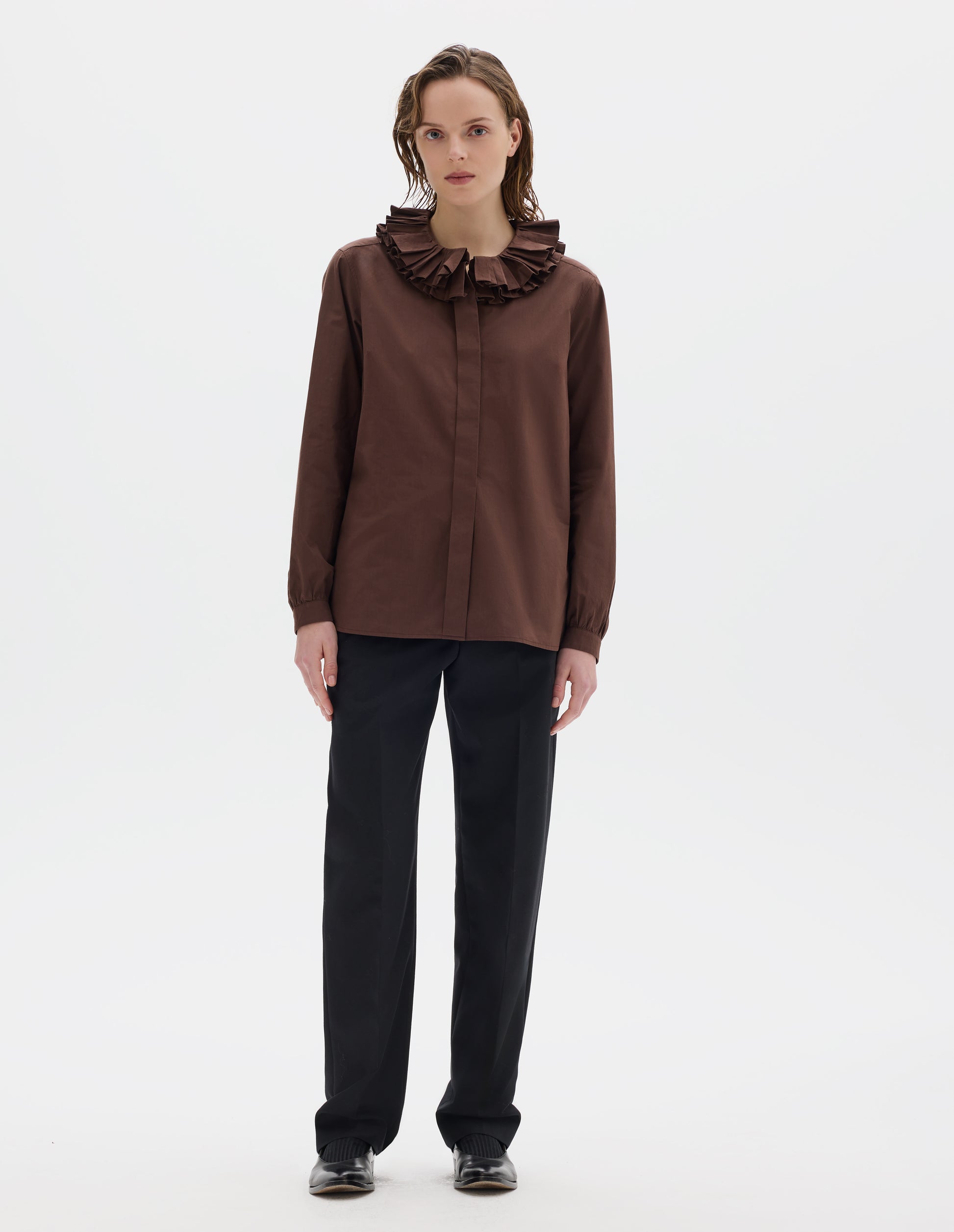 Full body image, front view of model wearing brown cotton shirt with double ruffle collar, concealed placket with mother of pearl buttons, bracelet-length sleeves with mother of pearl buttons on narrow, gathered cuffs, and with curved hem. Styled with black trousers and black leather shoes.
