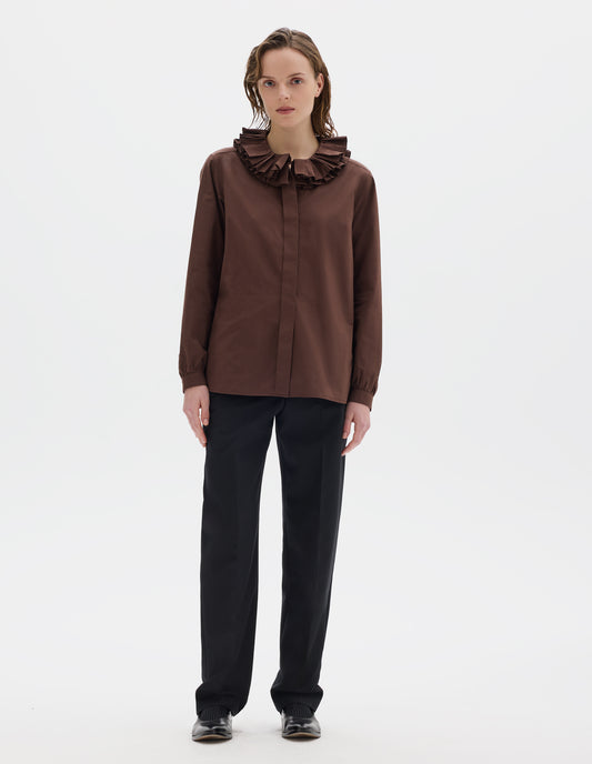 Full body image, front view of model wearing brown cotton shirt with double ruffle collar, concealed placket with mother of pearl buttons, bracelet-length sleeves with mother of pearl buttons on narrow, gathered cuffs, and with curved hem. Styled with black trousers and black leather shoes.