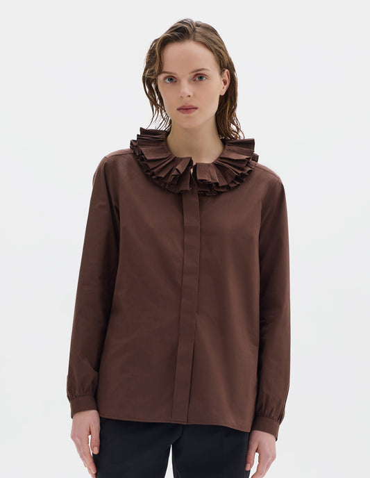 Cropped body image, front view of model wearing brown cotton shirt with double ruffle collar, concealed placket with mother of pearl buttons, bracelet-length sleeves with mother of pearl buttons on narrow, gathered cuffs, and with curved hem. Styled with black trousers.