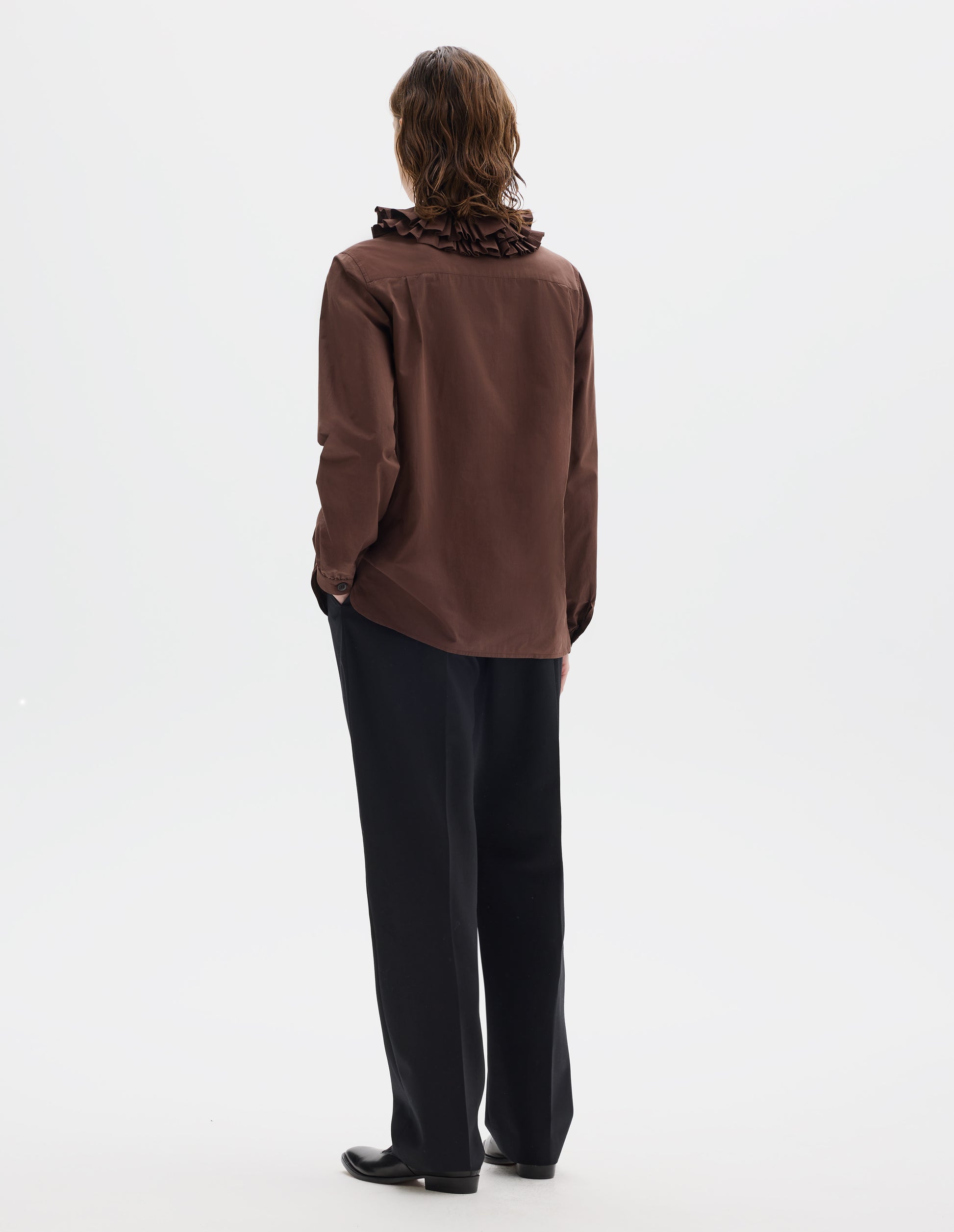 Full body image, back view of model wearing brown cotton shirt with double ruffle collar, bracelet-length sleeves with mother of pearl buttons on narrow, gathered cuffs, and with curved hem. Styled with black trousers and black leather shoes.