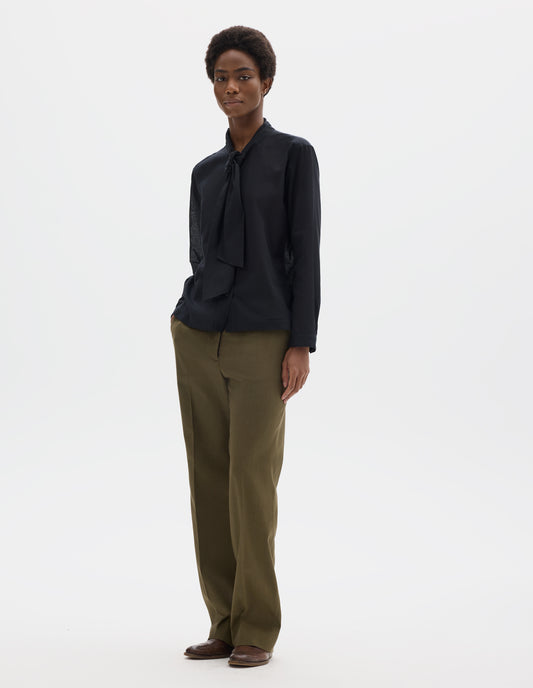 Full body image, front view of model wearing black cotton cashmere sheer shirt with attached loose tie, lipped placket, narrow cuffs and with square side split hem. Styled with khaki green trousers and brown leather brogues.