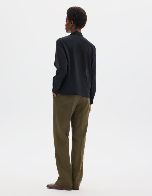 Full body image, back view of model wearing black cotton cashmere sheer shirt with mother of pearl buttons on narrow cuffs with pleats, and with square side split hem. Styled with khaki green trousers and brown leather brogues.