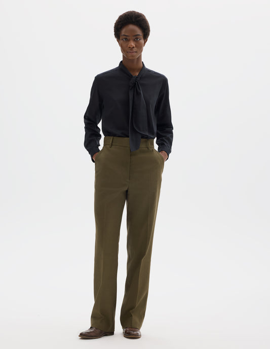 Full body image, front view of model wearing green cotton linen straight leg trousers with belt loops and slant pockets. Styled with black shirt and brown leather brogues.