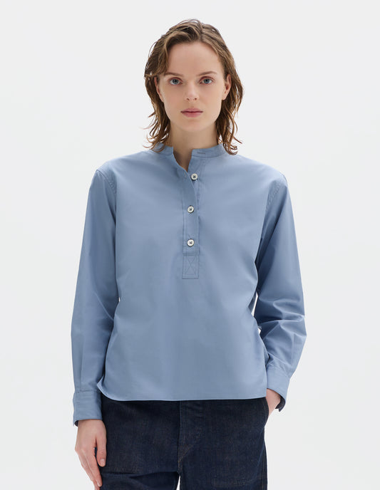 Cropped body image, front view of model wearing relaxed blue cotton poplin collarless pull on shirt with mother of pearl buttons on placket with box stitch detailing and stepped hem. Styled with dark blue jeans.
