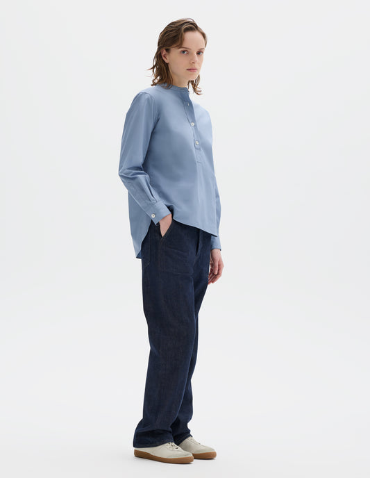Full body image, front view of model wearing relaxed blue cotton poplin collarless pull on shirt with mother of pearl buttons on placket with box stitch detailing and curved stepped hem. Styled with dark blue jeans and beige nubuck trainers.