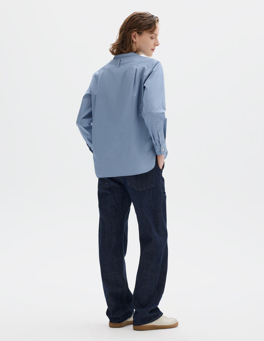 Full body image, back view of model wearing relaxed blue cotton poplin collarless shirt with narrow shoulder yoke with hanger loop, curved stepped hem and mother of pearl buttons on cuffs. Styled with dark blue jeans and beige nubuck trainers.
