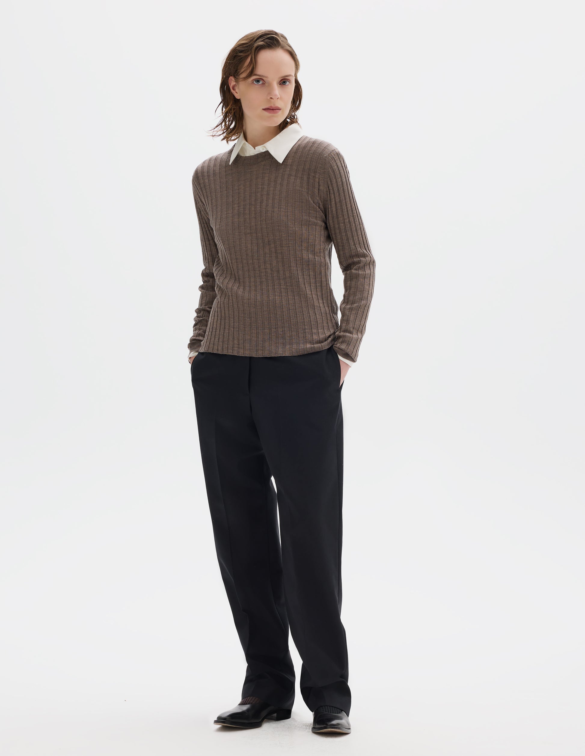 Full body image, front view of model wearing neat fitting dark beige merino top with wide rib through body, wide contrast rib on crew neck, and set-in long sleeves with fine rib cuffs. Styled over white shirt, and with black trousers and black leather shoes.