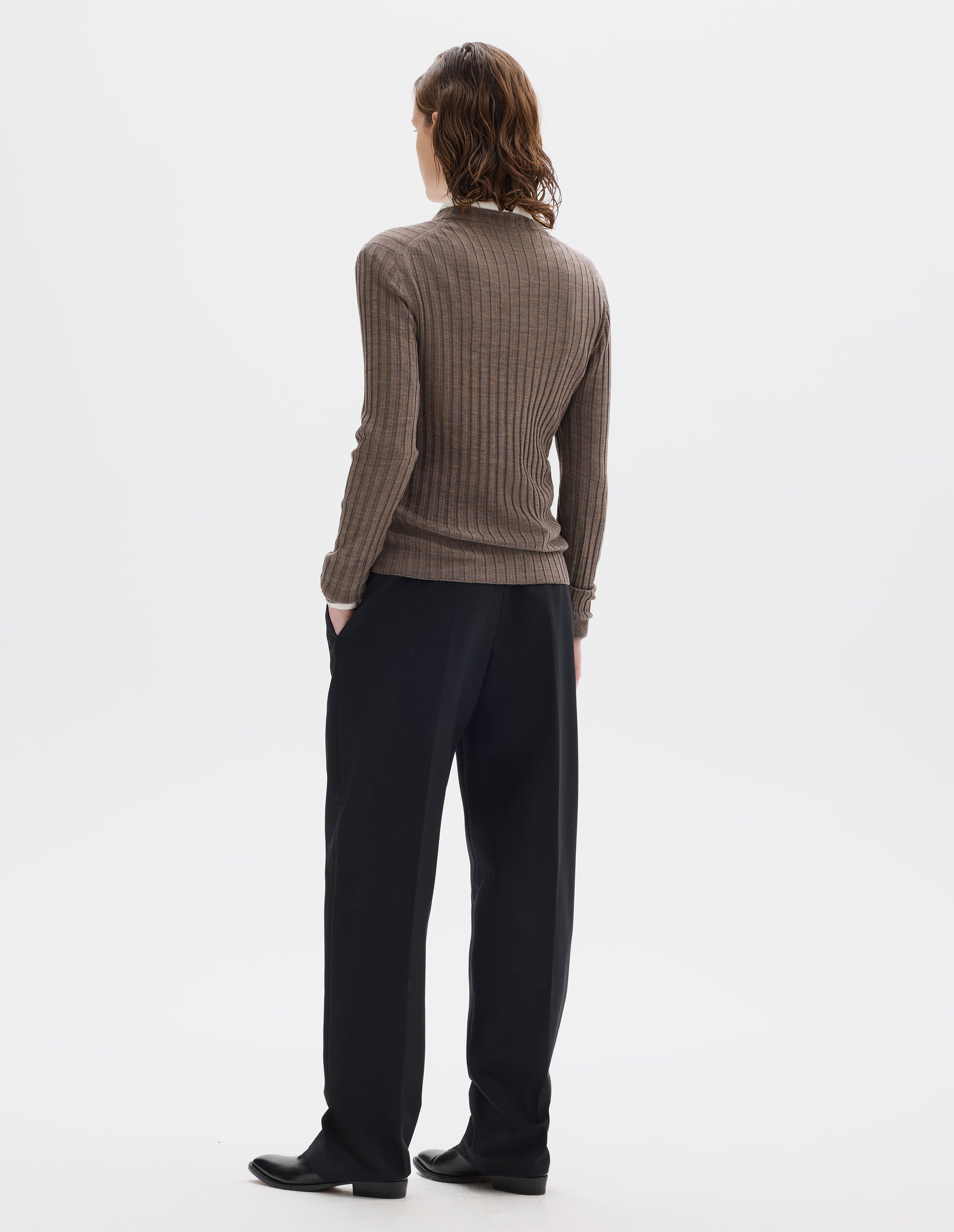 Full body image, back view of model wearing neat fitting dark beige merino top with wide rib through body, wide contrast rib on crew neck, and set-in long sleeves with fine rib cuffs. Styled over white shirt, and with black trousers and black leather shoes.