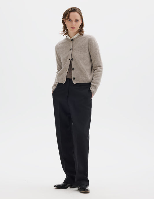 Full body image, front view of model wearing beige merino cashmere fully fashioned cardigan with dark horn buttons and with ribbed crew neck, cuffs and hem. Styled over beige shirt and dark beige jumper, and with black trousers and black leather shoes.