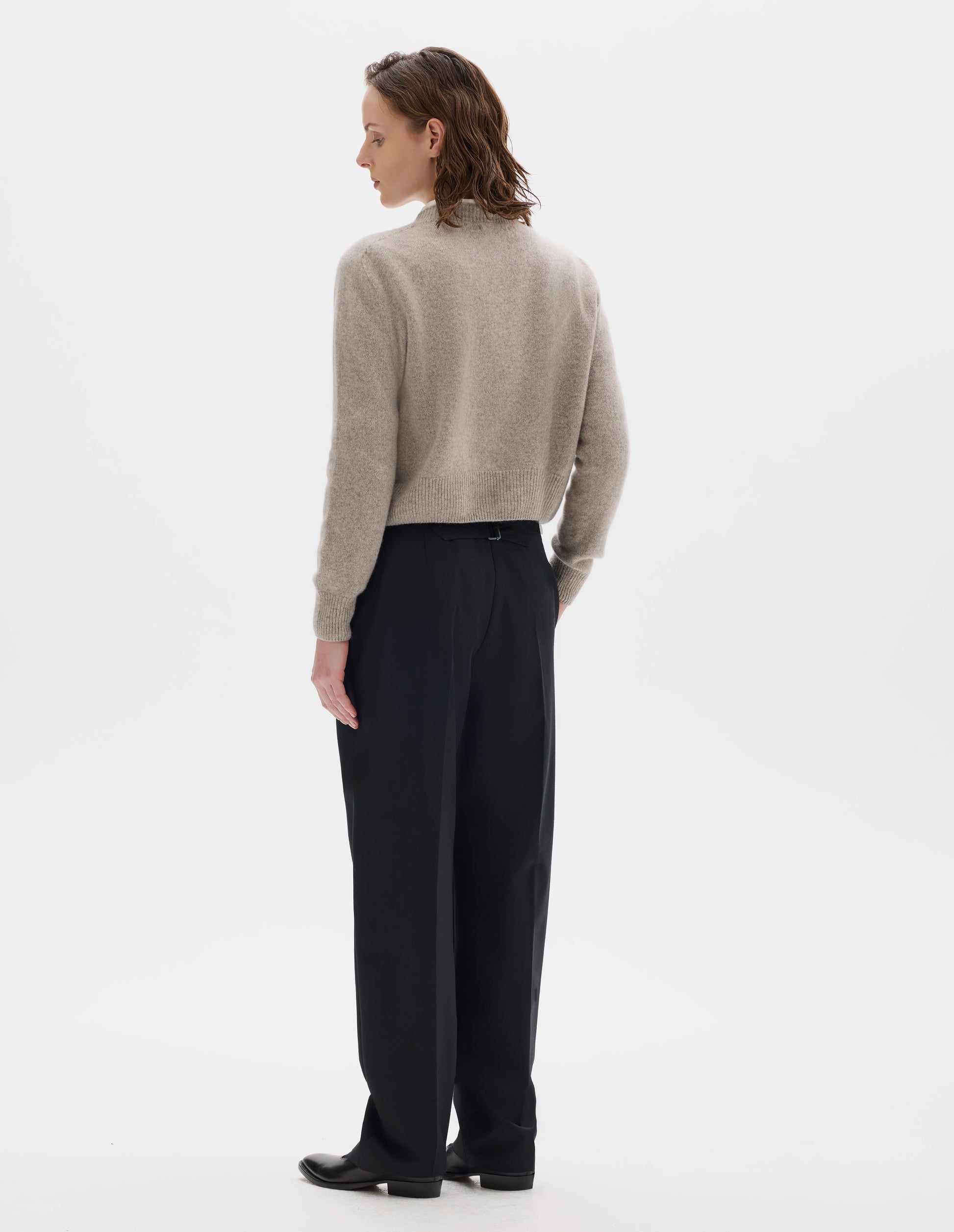 Full body image, back view of model wearing beige merino cashmere fully fashioned cardigan with ribbed crew neck, cuffs and hem. Styled over beige shirt, and with black trousers and black leather shoes.