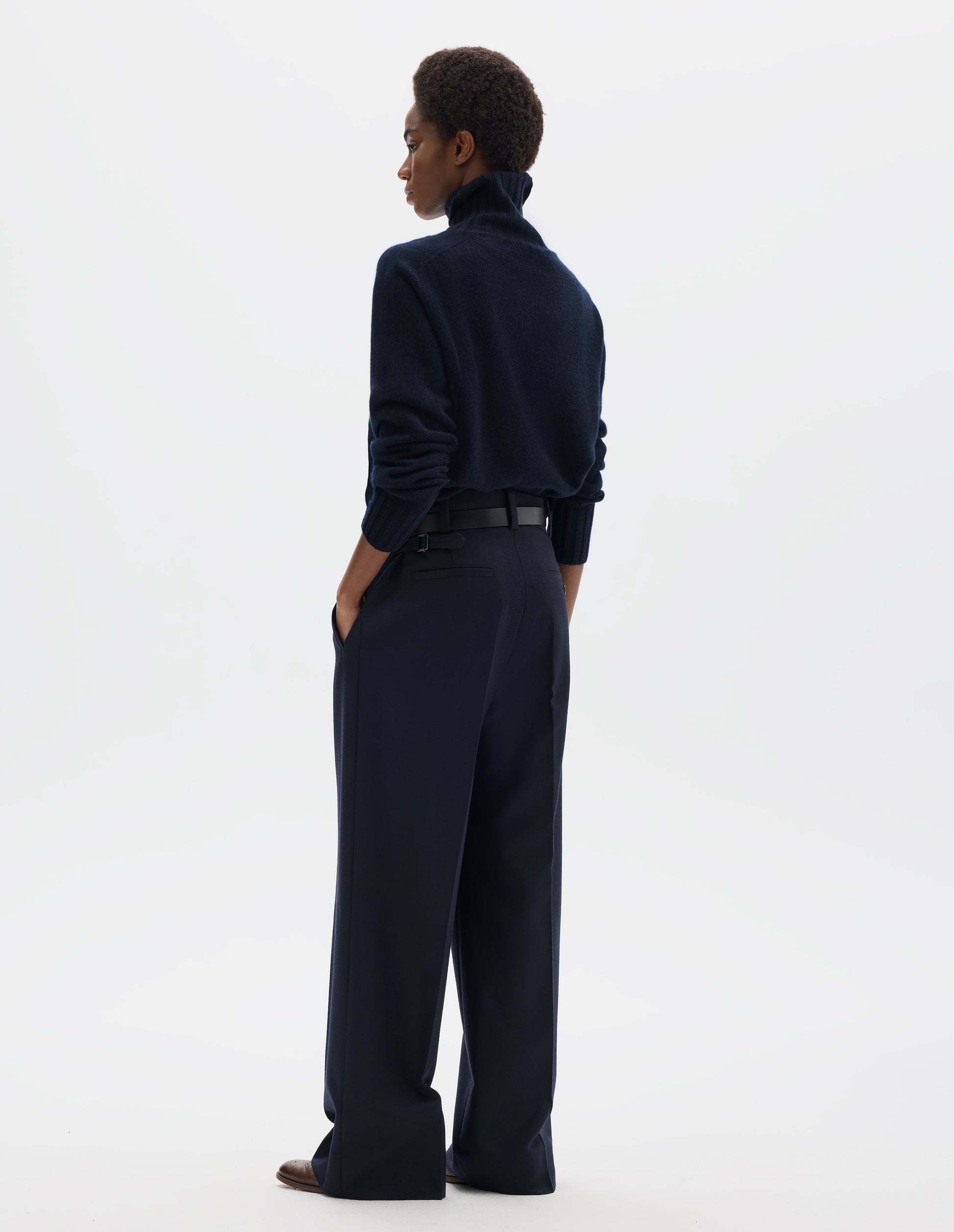 Full body image, back view of model wearing dark blue high waisted wool trousers with wide leg, belt loops on wide waistband, side adjusters and two welt back pockets, one with horn button fastening. Styled with dark blue roll neck jumper, black leather belt and brown leather brogues.