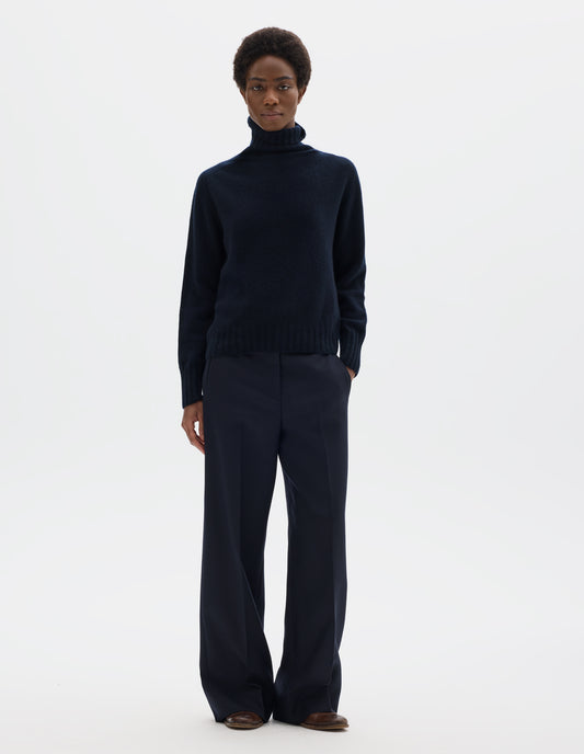 Full body image, front view of model wearing seamless dark blue cashmere roll neck jumper with contrast ribbing on neck, cuffs and hem. Styled with dark blue trousers and brown leather brogues.