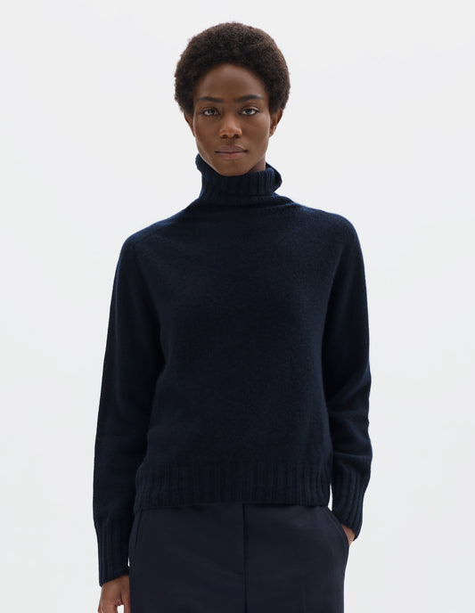 Cropped body image, front view of model wearing seamless dark blue cashmere roll neck jumper with contrast ribbing on neck, cuffs and hem. Styled with dark blue trousers.