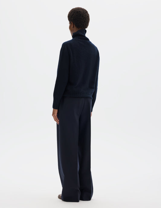 Full body image, back view of model wearing seamless dark blue cashmere roll neck jumper with contrast ribbing on neck, cuffs and hem. Styled with dark blue trousers and brown leather brogues.
