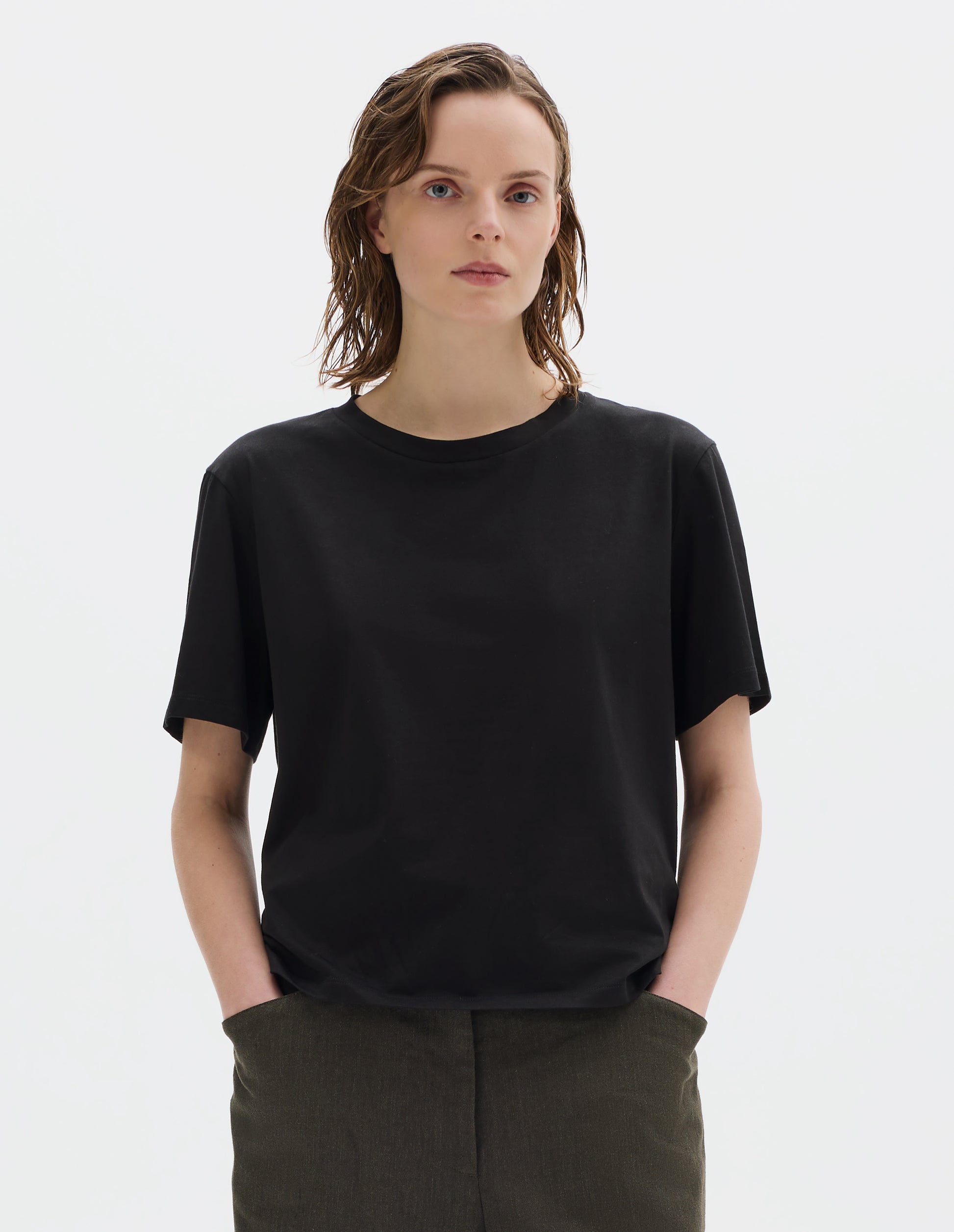 Cropped body image, front view of model wearing black relaxed cotton t-shirt with crew neck and boxy cut. Styled with brown trousers.