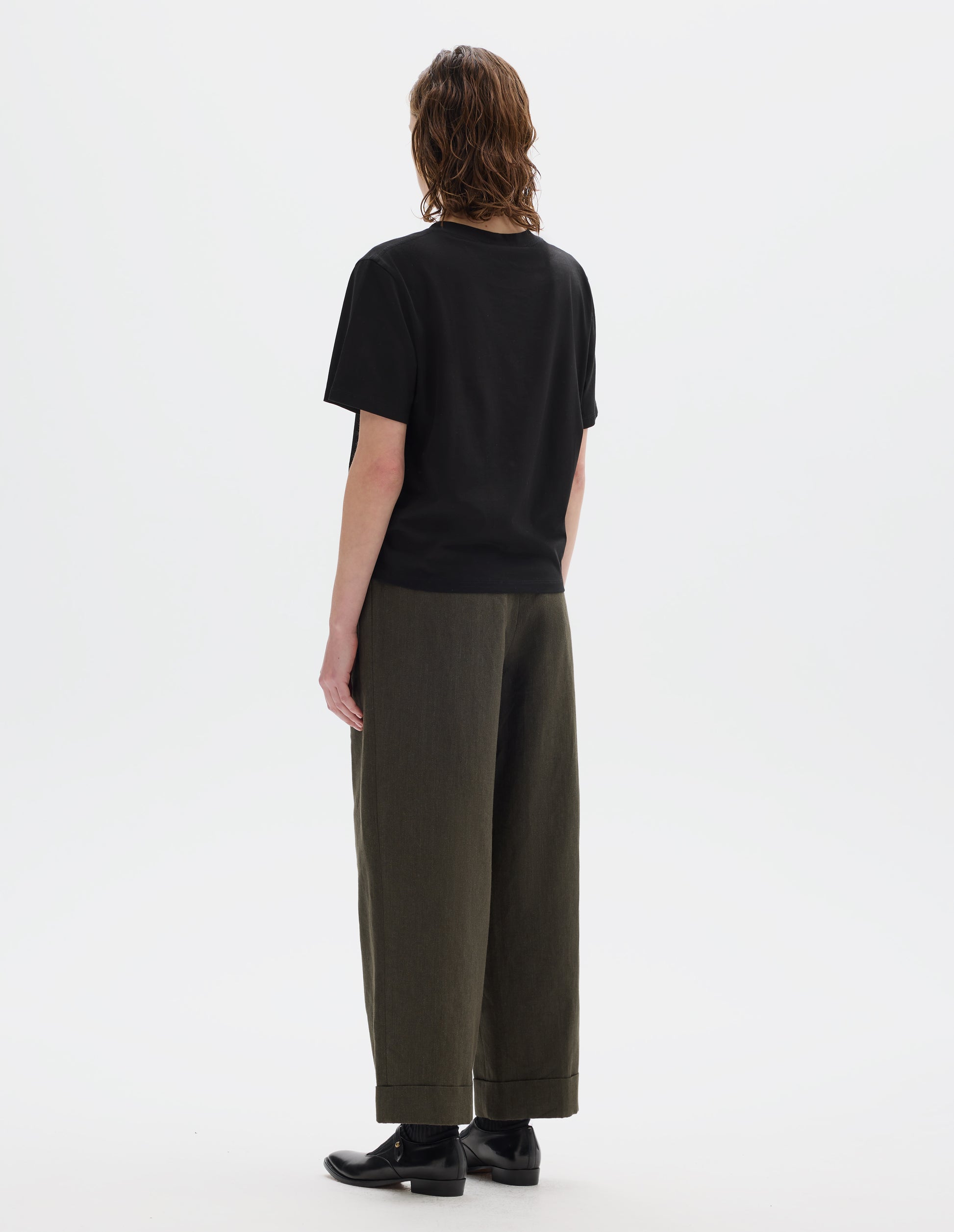 Full body image, back view of model wearing black relaxed cotton t-shirt with crew neck and boxy cut. Styled with brown trousers and black leather shoes.