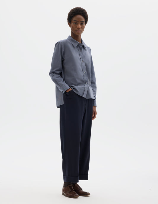 Full body image, front view of model wearing blue cotton shirt with mother of pearl buttons on lipped placket, stepped hem with curved front and straight-cut back. Styled with dark blue trousers and brown leather brogues.