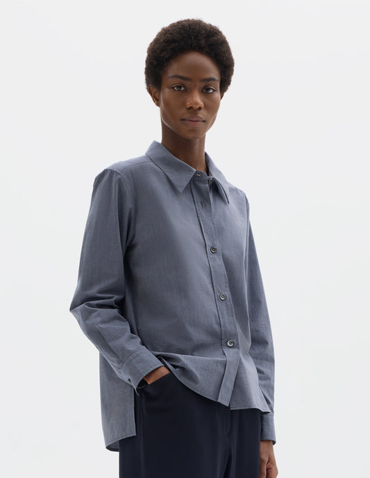 Cropped body image, front view of model wearing blue cotton shirt with mother of pearl buttons on lipped placket, stepped hem with curved front and straight-cut back. Styled with dark blue trousers.