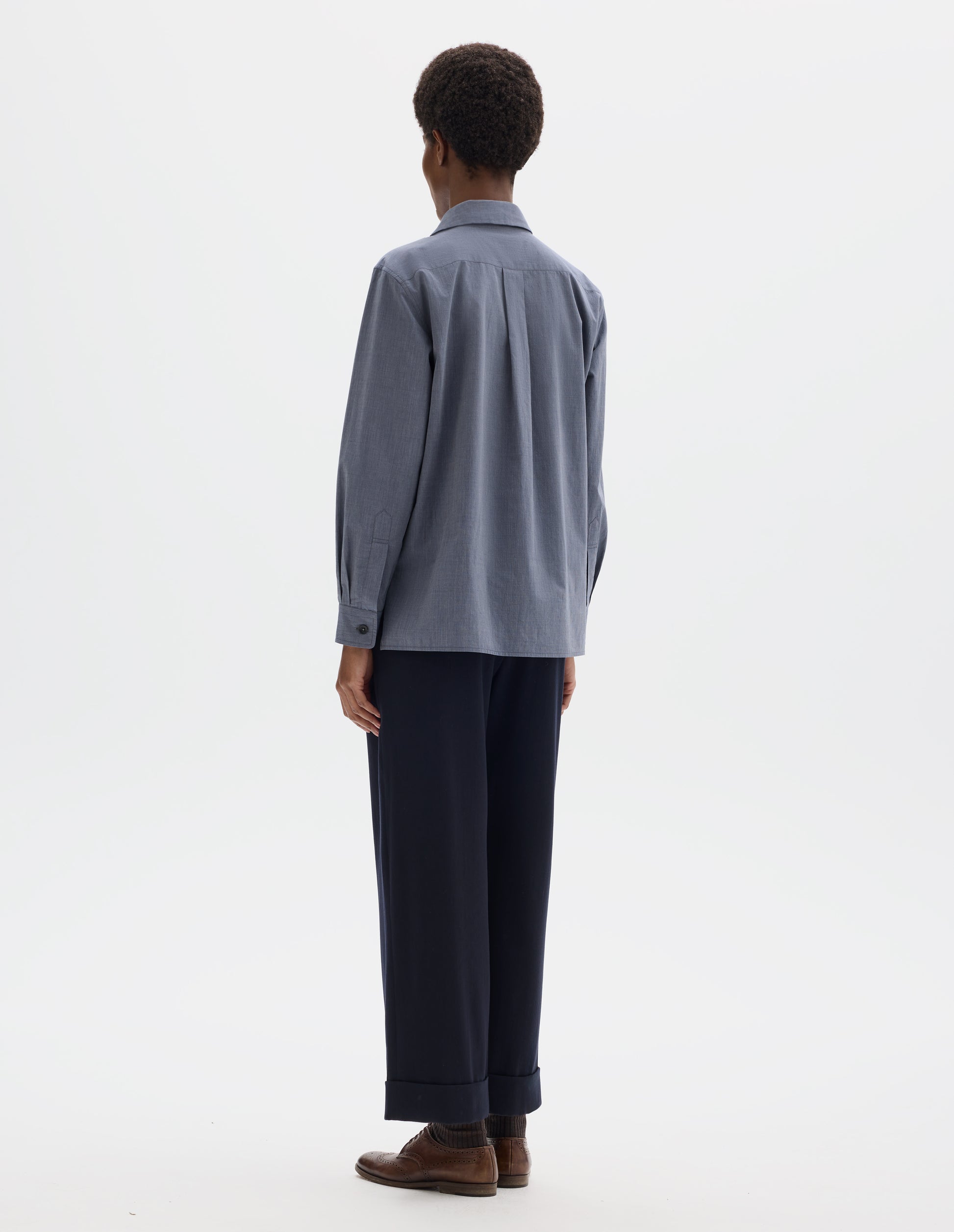 Full body image, back view of model wearing blue cotton shirt with pleat on back yoke, mother of pearl buttons cuffs and stepped hem. Styled with dark blue trousers and brown leather brogues.