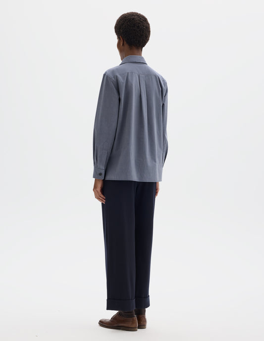 Full body image, back view of model wearing blue cotton shirt with pleat on back yoke, mother of pearl buttons cuffs and stepped hem. Styled with dark blue trousers and brown leather brogues.