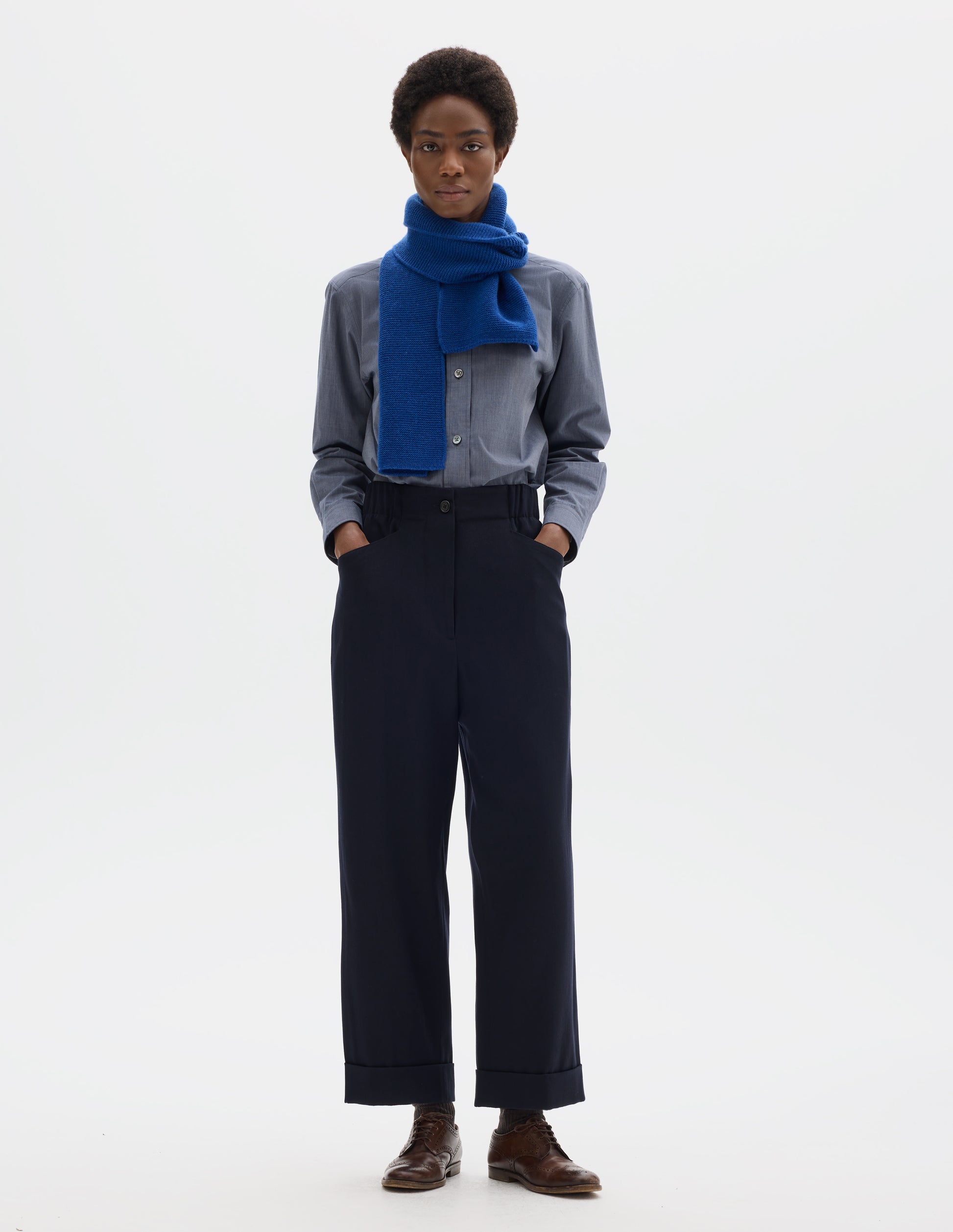 Full body image, front view of model wearing relaxed dark blue cropped wool trousers with zip fly and horn button fastening on elasticated waistband, frog pockets and stitch-down turn-up hem. Styled with blue shirt, bright blue scarf, dark brown socks and brown leather brogues.