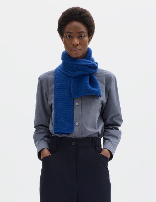 Cropped image, front view of female model wearing blue merino cashmere scarf knitted in garter stitch. Styled with blue shirt and dark blue trousers.