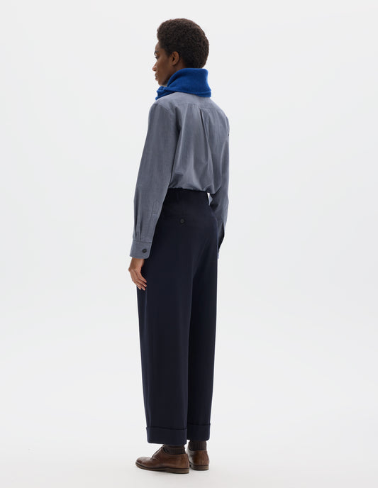 Full body image, back view of model wearing relaxed dark blue cropped wool trousers with elasticated waistband, frog pockets, horn button-through jet back pockets and stitch-down turn-up hem. Styled with blue shirt, bright blue scarf, dark brown socks and brown leather brogues.