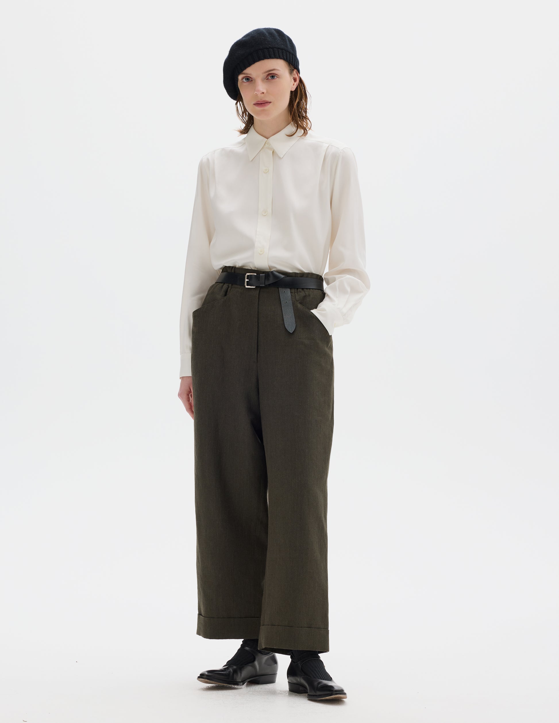 Full body image, front view of model wearing relaxed dark green cropped linen trousers with zip fly and horn button fastening on elasticated waistband, frog pockets and stitch-down turn-up hem. Styled with black knitted beret, white shirt, black leather belt, black socks and black leather shoes.