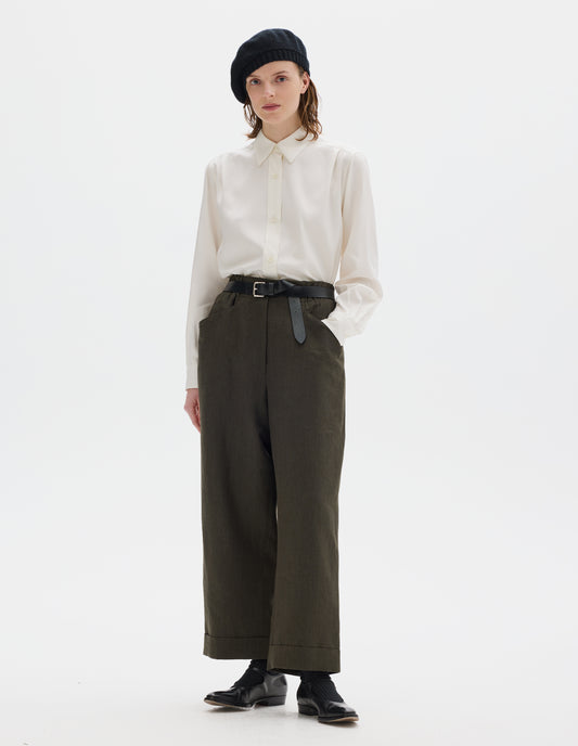 Full body image, front view of model wearing relaxed dark green cropped linen trousers with zip fly and horn button fastening on elasticated waistband, frog pockets and stitch-down turn-up hem. Styled with black knitted beret, white shirt, black leather belt, black socks and black leather shoes.
