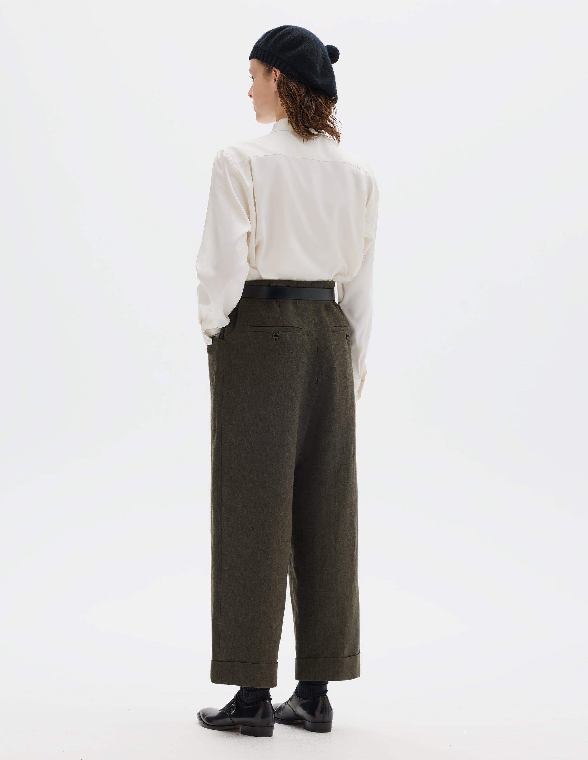Full body image, back view of model wearing relaxed dark green cropped linen trousers with elasticated waistband, frog pockets, horn button-through jet back pockets and stitch-down turn-up hem. Styled with black knitted beret, white shirt, black leather belt, black socks and black leather shoes.