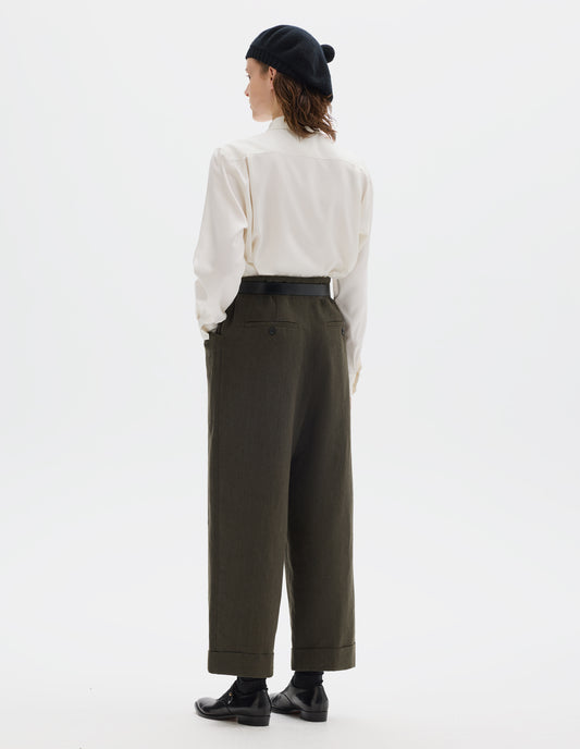 Full body image, back view of model wearing relaxed dark green cropped linen trousers with elasticated waistband, frog pockets, horn button-through jet back pockets and stitch-down turn-up hem. Styled with black knitted beret, white shirt, black leather belt, black socks and black leather shoes.