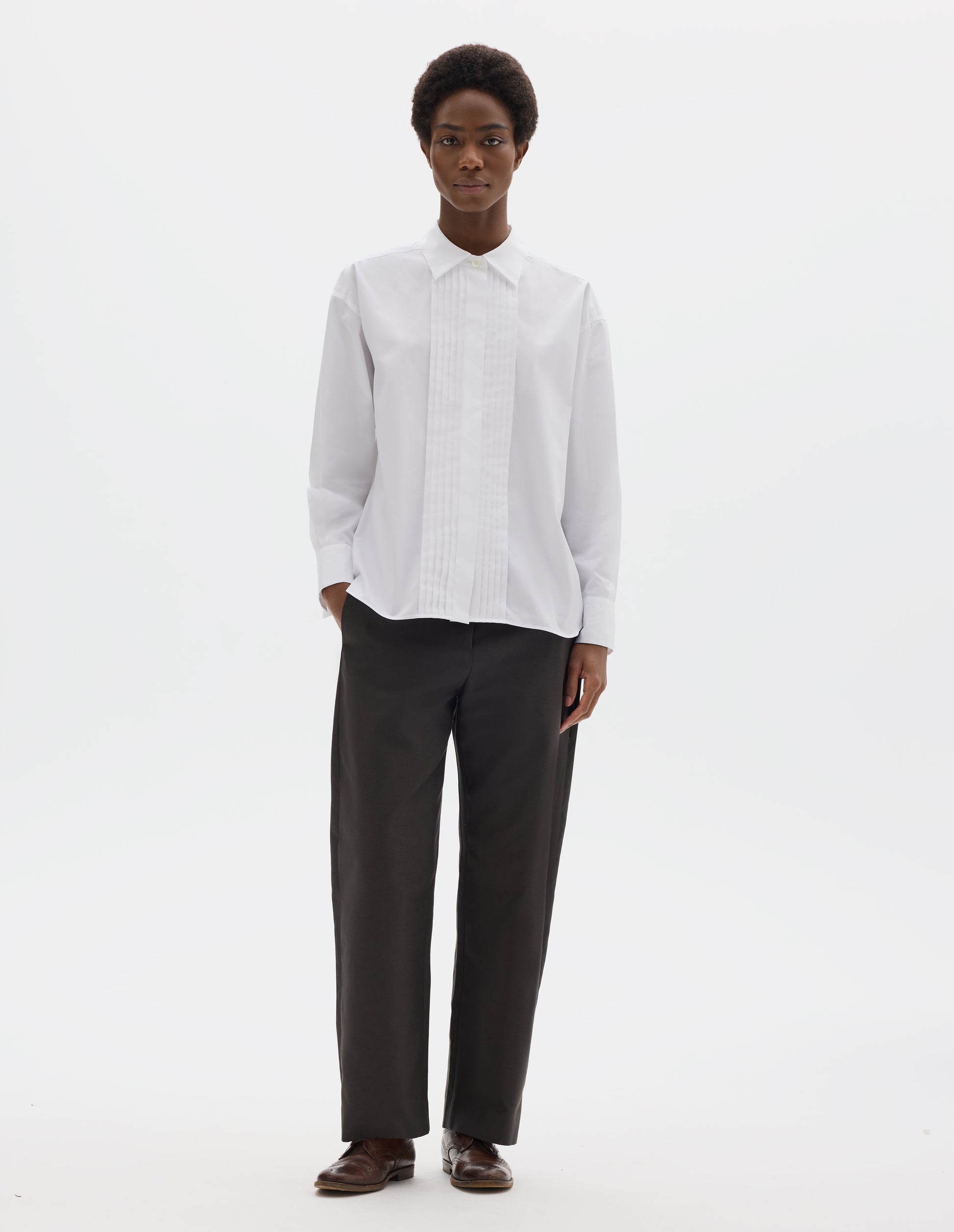 Full body image, front view of model wearing white oversized cotton shirt with mother of pearl button closure, concealed placket and pin tuck detailing at front, and with curved hem. Styled with dark grey trousers and brown leather brogues.