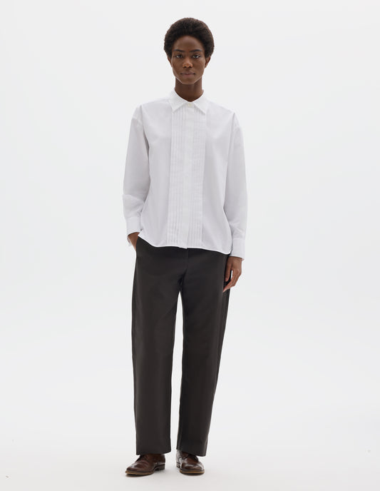 Full body image, front view of model wearing white oversized cotton shirt with mother of pearl button closure, concealed placket and pin tuck detailing at front, and with curved hem. Styled with dark grey trousers and brown leather brogues.