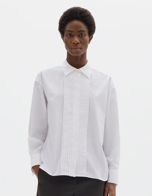 Cropped body image, front view of model wearing white oversized cotton shirt with mother of pearl button closure, concealed placket and pin tuck detailing at front, and with curved hem. Styled with dark grey trousers.