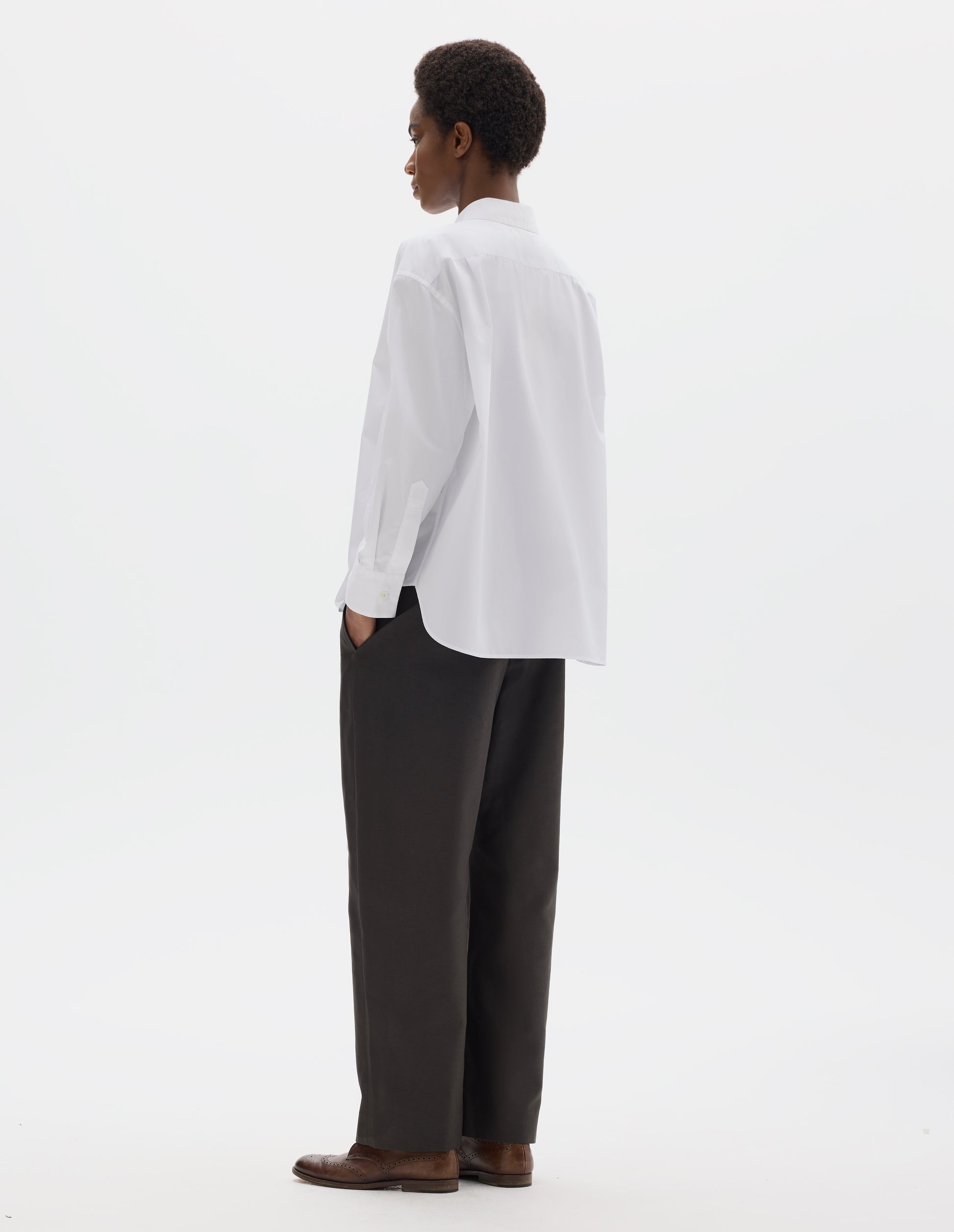 Full body image, back view of model wearing white oversized cotton shirt with back yoke and two small pleats, mother of pearl buttons on cuffs, and curved hem. Styled with dark grey trousers and brown leather brogues.