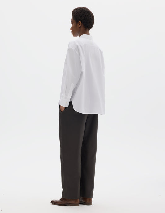 Full body image, back view of model wearing white oversized cotton shirt with back yoke and two small pleats, mother of pearl buttons on cuffs, and curved hem. Styled with dark grey trousers and brown leather brogues.