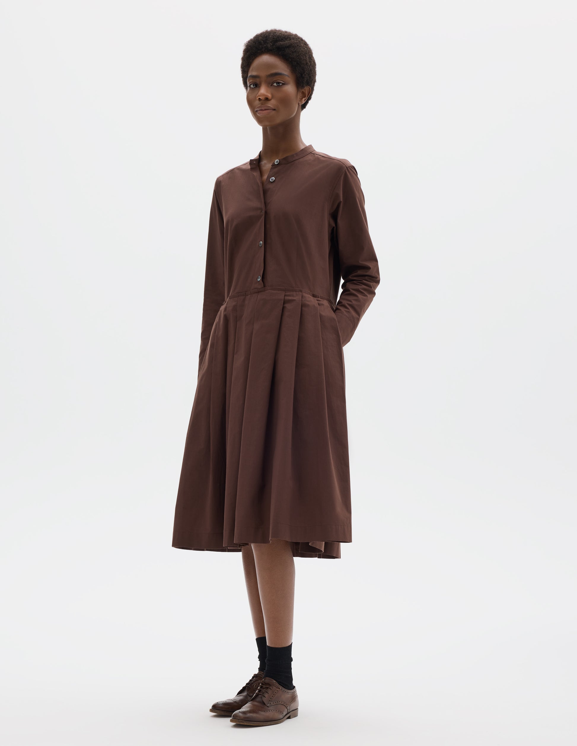 Full body image, front view of model wearing relaxed brown collarless cotton midi dress with mother of pearl buttons on half placket, long sleeves with shirt cuffs, and pleated skirt with concealed side pockets. Styled with black socks and brown leather brogues.