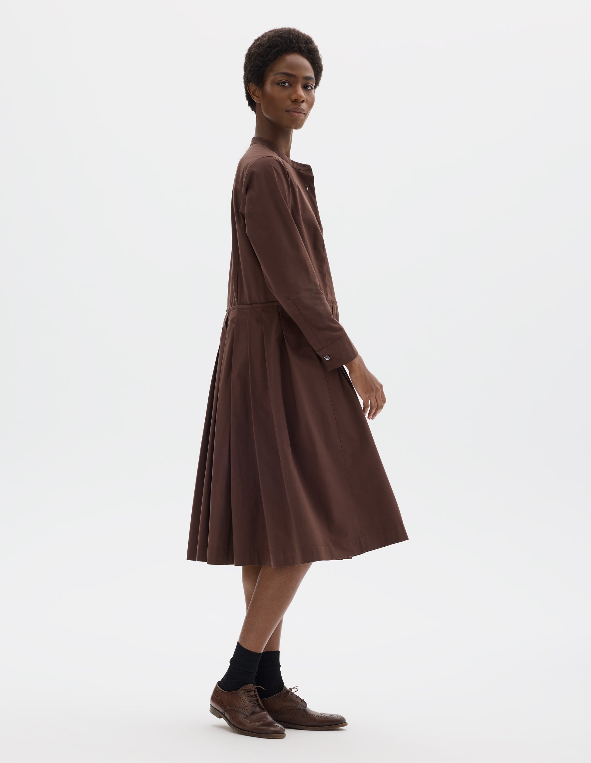 Full body image, side view of model wearing relaxed brown collarless cotton midi dress with shirt yoke, with mother of pearl buttons on half placket, long sleeves with mother of pearl buttons on cuffs and pleated skirt with concealed side pockets. Styled with black socks and brown leather brogues.