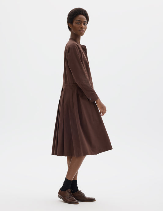 Full body image, side view of model wearing relaxed brown collarless cotton midi dress with shirt yoke, with mother of pearl buttons on half placket, long sleeves with mother of pearl buttons on cuffs and pleated skirt with concealed side pockets. Styled with black socks and brown leather brogues.