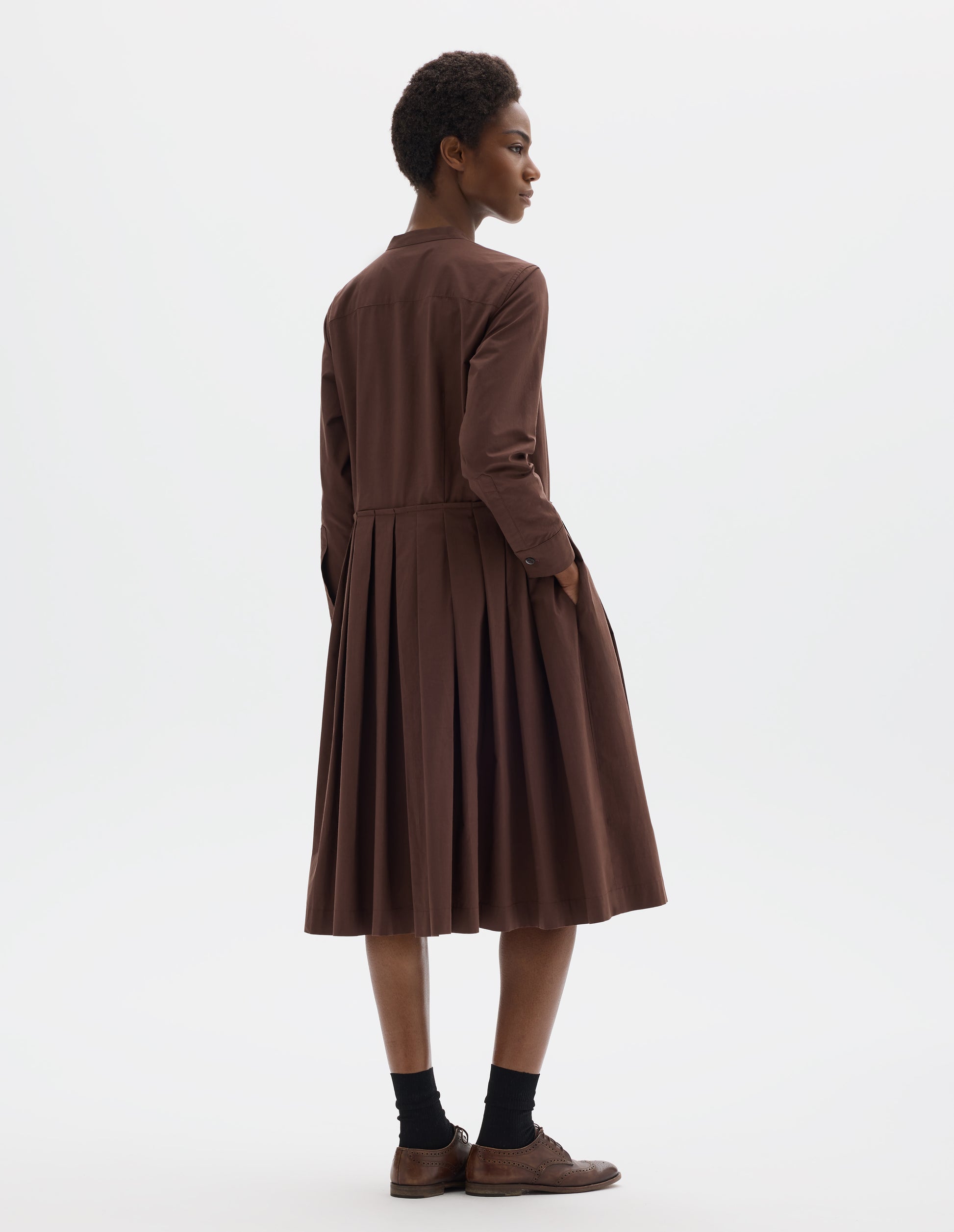Full body image, back view of model wearing relaxed brown collarless cotton midi dress with shirt yoke, long sleeves with mother of pearl buttons on cuffs and pleated skirt with concealed side pockets. Styled with black socks and brown leather brogues.