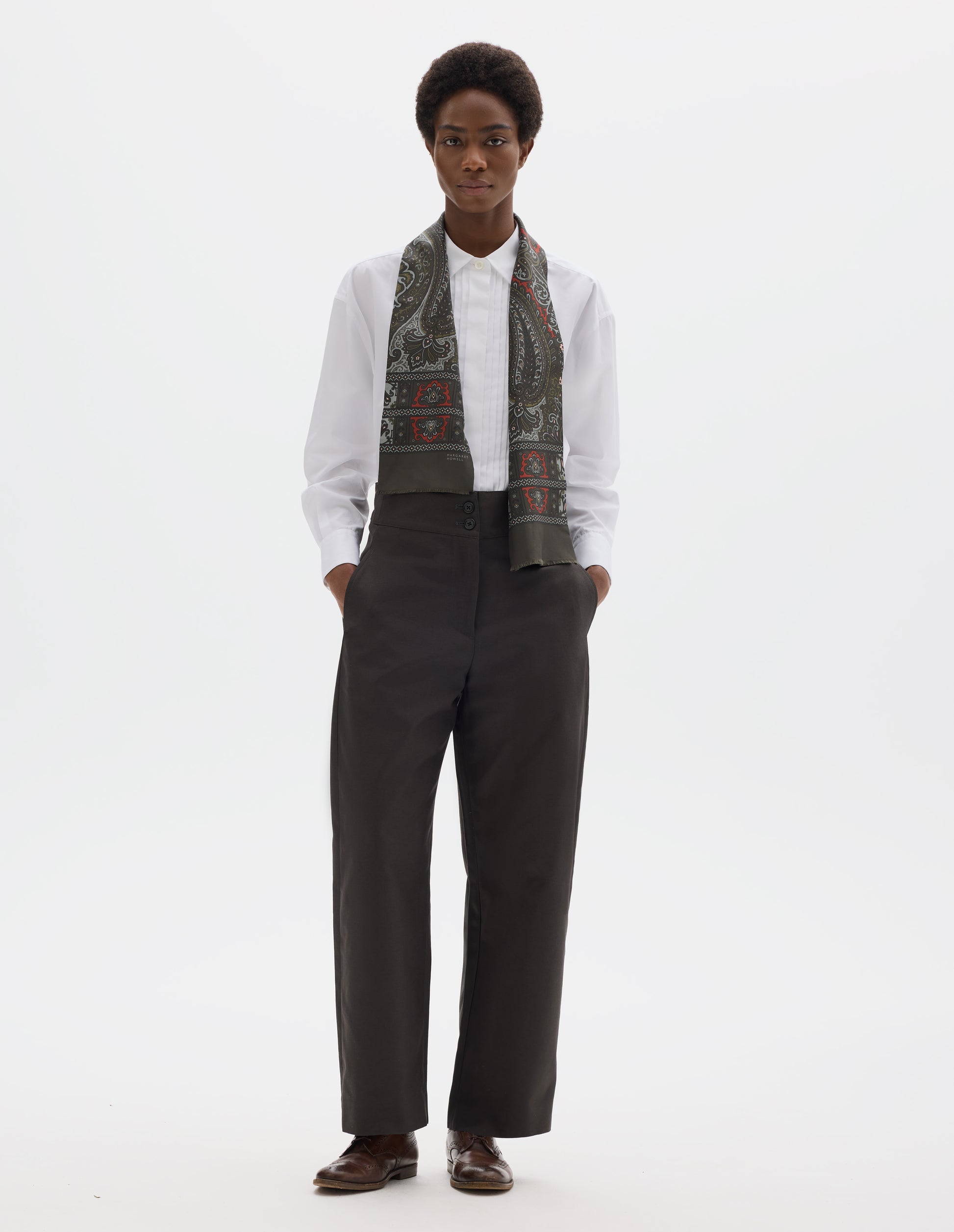 Full body image, front view of model wearing high waisted grey cotton trousers with double horn button fastening and concealed side pockets with stitch detailing. Styled with white shirt, dark green silk scarf and brown leather brogues.