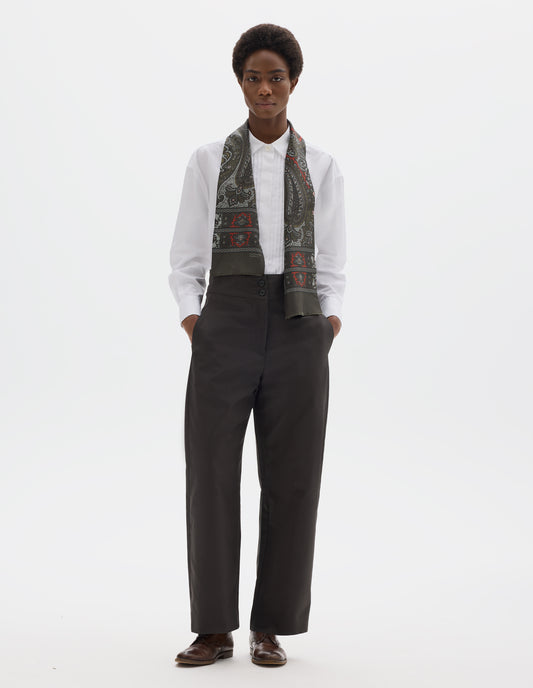 Full body image, front view of model wearing high waisted grey cotton trousers with double horn button fastening and concealed side pockets with stitch detailing. Styled with white shirt, dark green silk scarf and brown leather brogues.