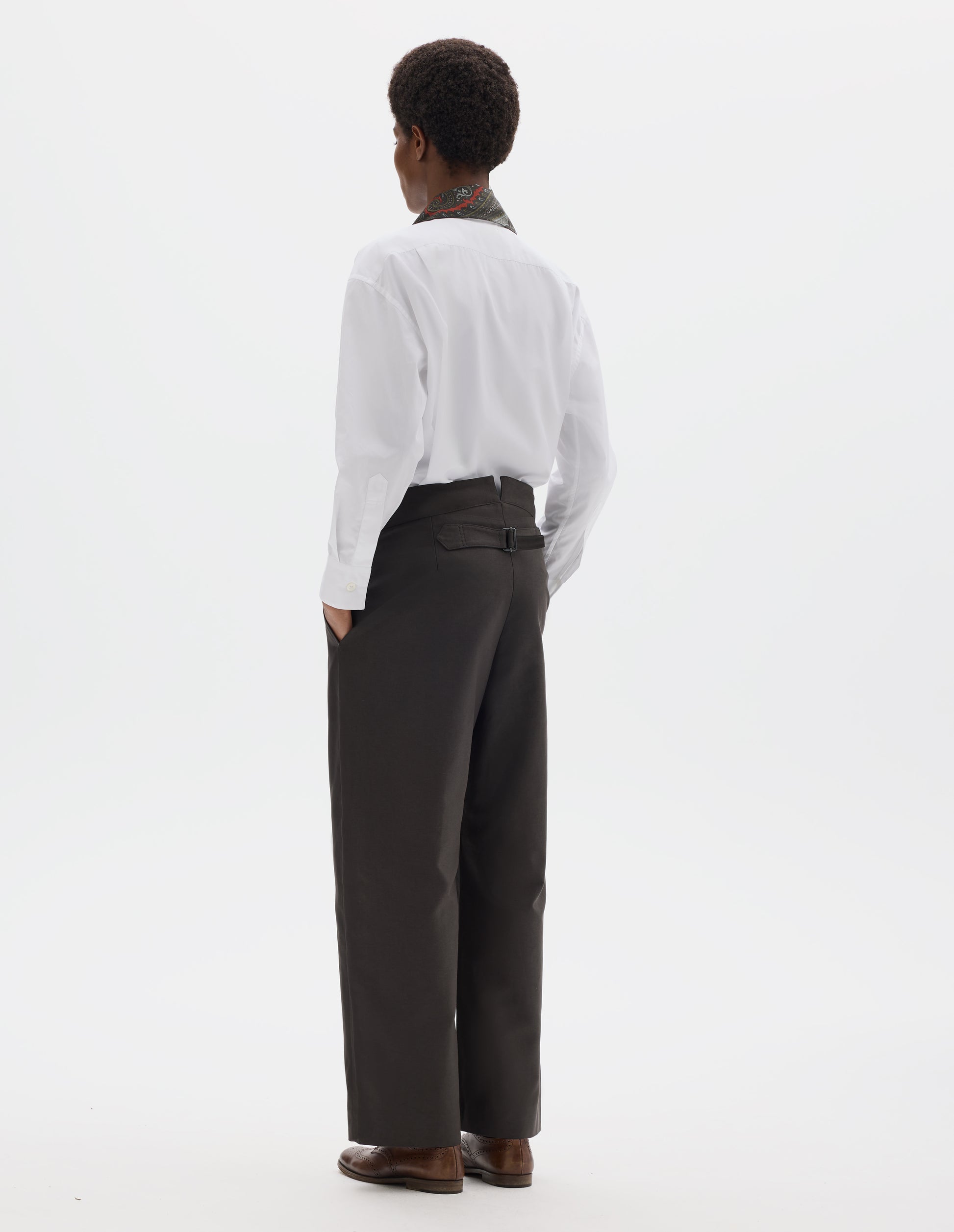 Full body image, back view of model wearing high waisted grey cotton trousers with back cinch. Styled with white shirt, dark green silk scarf and brown leather brogues.