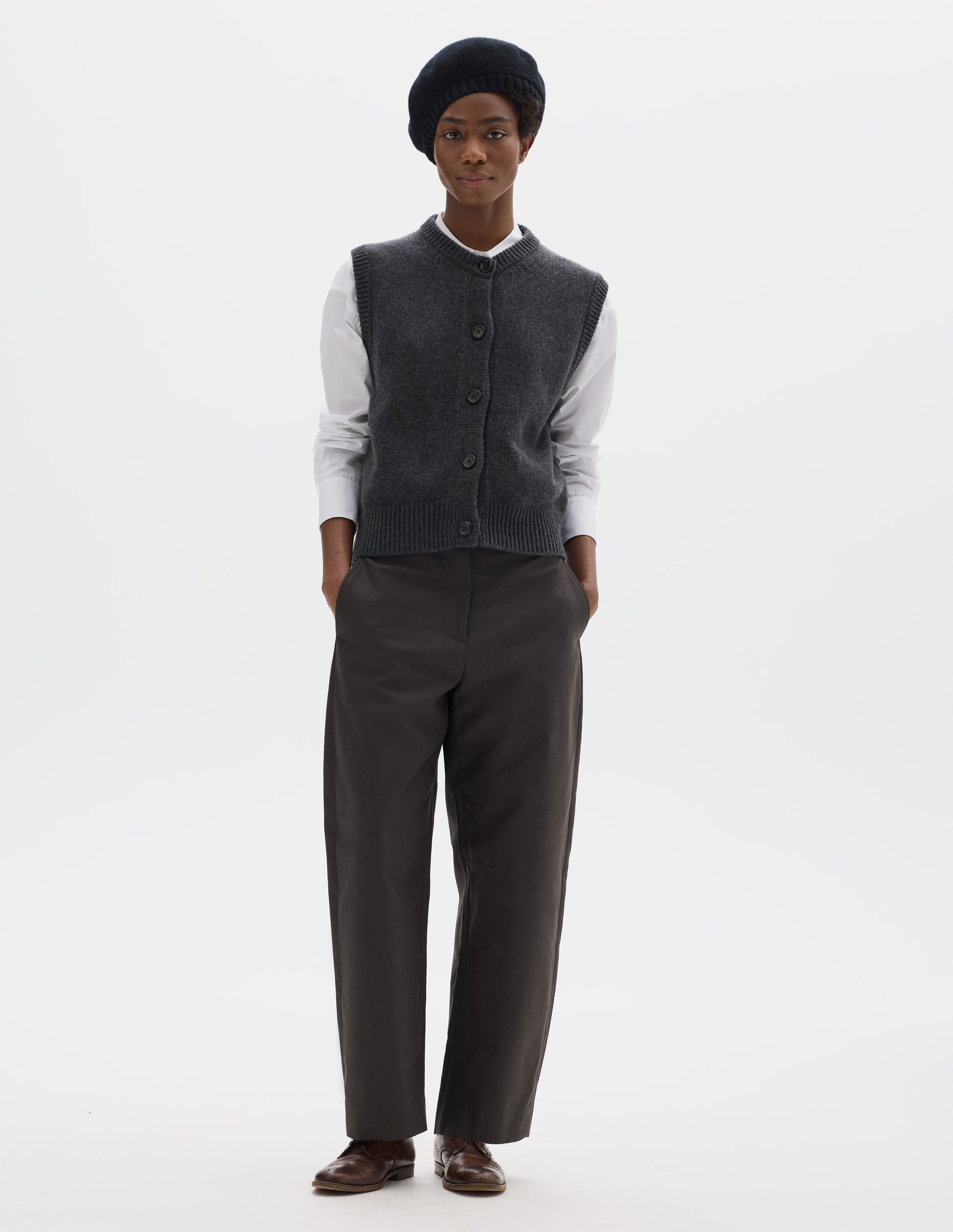 Full body image, front view of model wearing relaxed dark grey cashmere merino knitted gilet with horn buttons on Milano stitch placket, rib detailing at neck, armholes and hem. Styled over white shirt, and with black beret, grey trousers and brown leather brogues. 