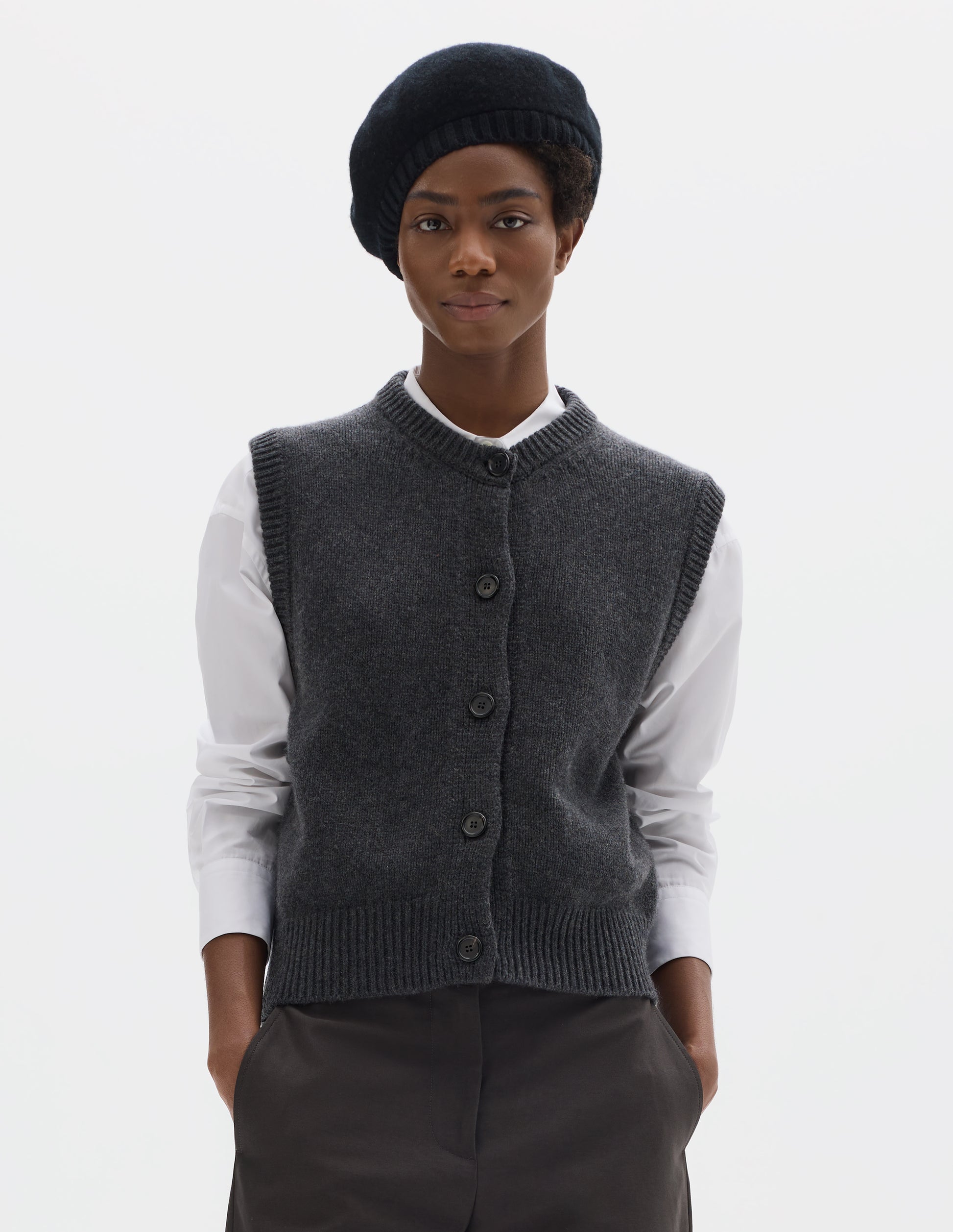 Cropped body image, front view of model wearing relaxed dark grey cashmere merino knitted gilet with horn buttons on Milano stitch placket, rib detailing at neck, armholes and hem. Styled over white shirt, and with black beret and grey trousers.